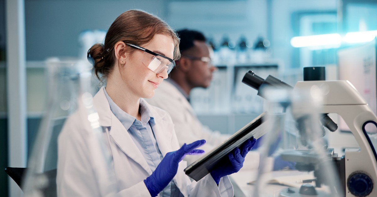 A scientist at the microscope using a tablet to enter data. She wears blue latex gloves, a white lab coat, and glasses.