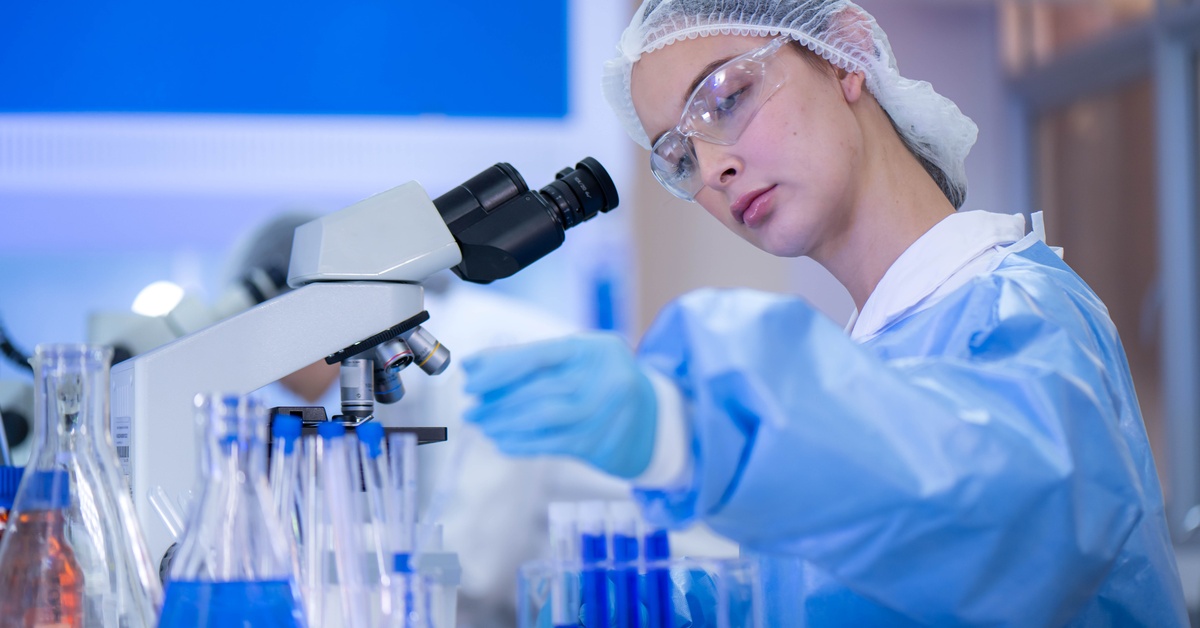 A lab specialist in a blue PPE gown and hair net works at their station with test tubes and a microscope.