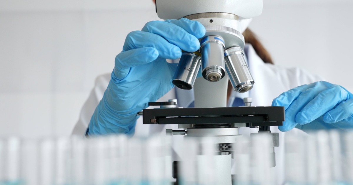 A scientist in a white lab coat and blue latex gloves looks through a microscope as test tubes sit in front of it.