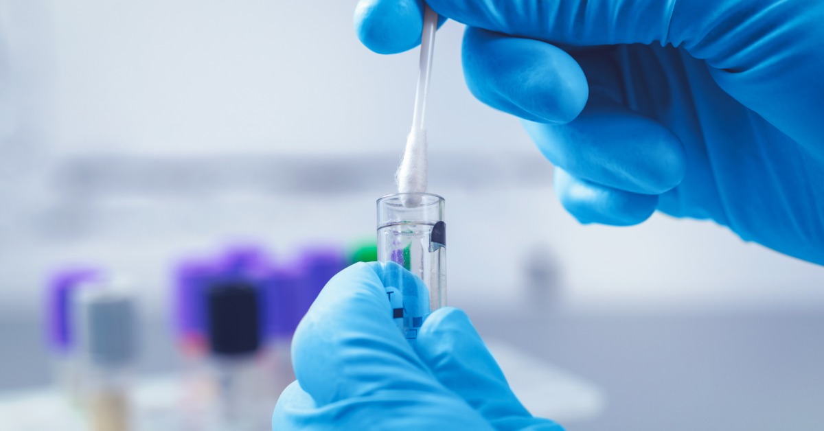A close-up of a professional in blue latex gloves. They hold a test tube with a Q-tip to test the substance.